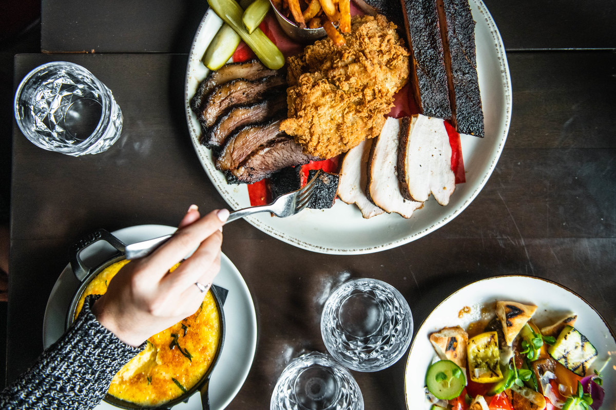 A Carbon Bar BBQ platter with sliced brisket, fried chicken, smoked meats, cornbread, and a hand reaching in with a fork
